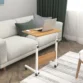 Oak wood bed table with a white metal frame, adjustable height, and rolling wheels, holding a laptop, ideal for use on a couch or bed.