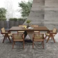 Outdoor view of a rectangular wooden dining table with six cane-back armchairs arranged on a textured rug.