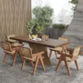 Angled shot showing the wooden dining table with sculptural crossed base and cane armchairs around it.