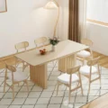 Light wood rectangular extendable table with slatted pedestal base, paired with six cane-back chairs with white seats in a bright modern dining room.