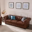 Brown Chesterfield sofa with tufted back, rolled arms, and nailhead trim, styled with gray and blue cushions beneath three framed artworks in a bright living room.
