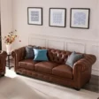 Brown Chesterfield sofa with tufted back, rolled arms, and nailhead trim, styled with gray and blue cushions beneath three framed artworks in a bright living room.