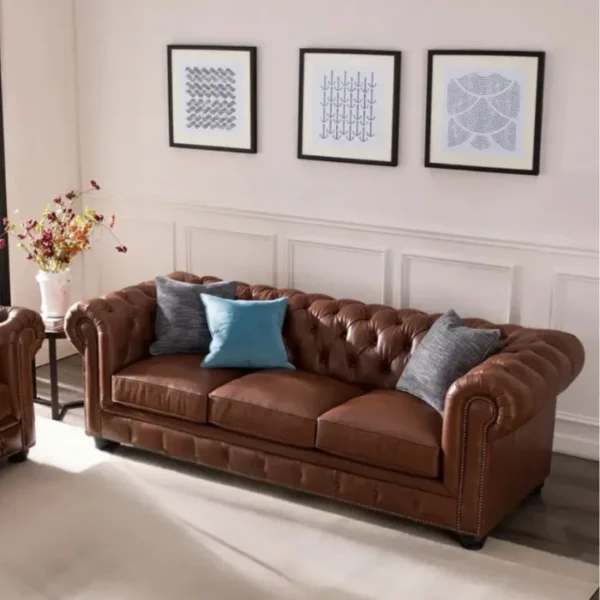 Brown Chesterfield sofa with tufted back, rolled arms, and nailhead trim, styled with gray and blue cushions beneath three framed artworks in a bright living room.