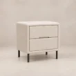 Angled view of Nolven Softline matching two-drawer bedside table in warm ivory upholstery with slim black metal legs.